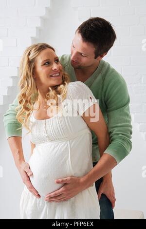Man standing behind his pregnant partner holding her belly with his hands as she smiles and looks up at him Stock Photo