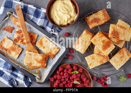 freshly baked portions of puff pastry on a baking sheet to cook french dessert millefeuille, fresh ripe raspberry on a plate and cream in a bowl on a  Stock Photo