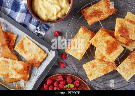 freshly baked portions of puff pastry on a baking sheet to cook french dessert millefeuille, fresh ripe raspberry on a plate and cream in a bowl on a  Stock Photo