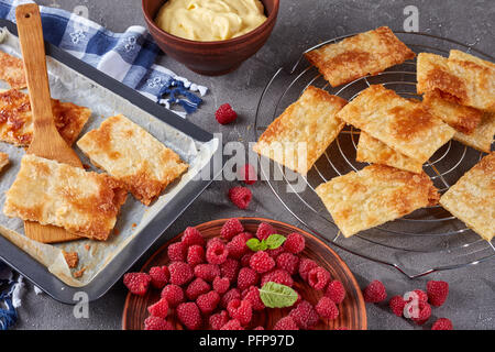 freshly baked portions of puff pastry on a baking sheet to cook french dessert millefeuille, fresh ripe raspberry on a plate and cream in a bowl on a  Stock Photo