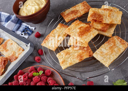 freshly baked portions of puff pastry on a baking sheet to cook french dessert millefeuille, fresh ripe raspberry on a plate and cream in a bowl on a  Stock Photo