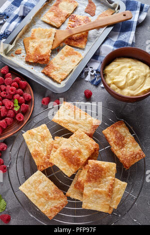 freshly baked portions of puff pastry on a baking sheet to cook french dessert millefeuille, fresh ripe raspberry on a plate and cream in a bowl on a  Stock Photo