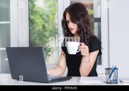 Beautiful young girl working on laptop at office desk and drinking cup of coffee. Modern business and job concept. Close up, selective focus Stock Photo