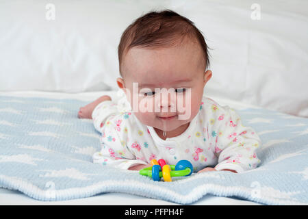 Baby girl lying on front on blue blanket on bed drooling looking at teething ring Stock Photo