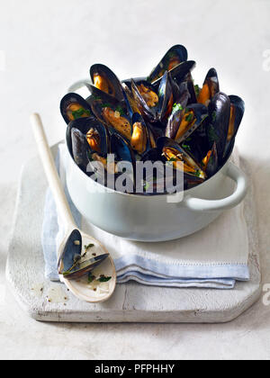 Moules marinieres served in bowl on chopping board with wooden spoon Stock Photo