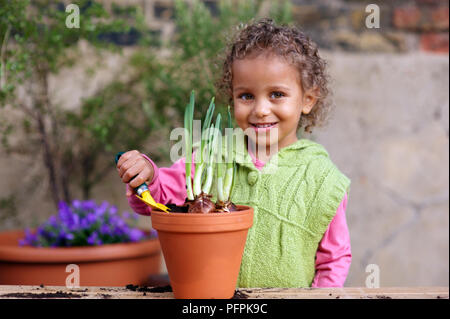 Girl (3.5 years) holding trowel next to flowerpot containing daffodil bulbs Stock Photo