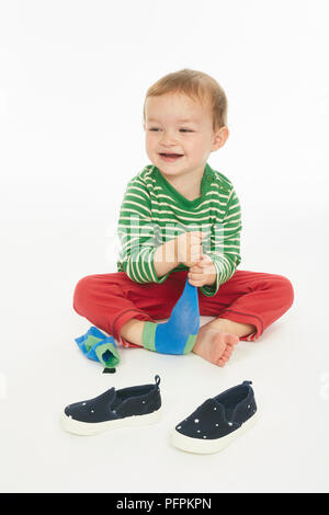 Little boy in green stripey top sitting, taking off shoes and socks  (Model age - 22 months) Stock Photo