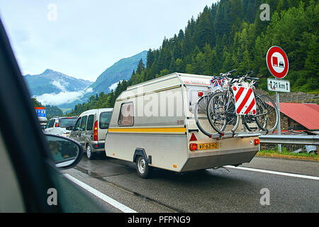 Caravan or travel trailer with hanging bicycles attached to a car on the alpine road in Swiss alps. Stock Photo