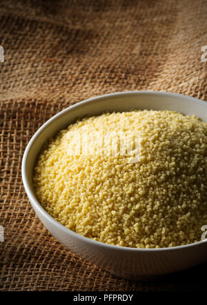 Heap of raw, uncooked couscous in white bowl on burlap background with selective focus Stock Photo