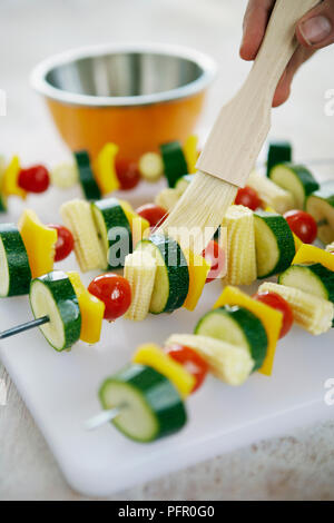 Using basting brush to apply sunflower oil to raw vegetable kebabs on skewers on plastic chopping board in kitchen Stock Photo