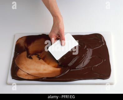 Man using plastic scraper to spread melted dark chocolate on chopping board, close-up Stock Photo