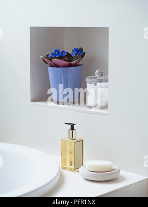 Blue flowers in plant pot and cotton wool ball in jar in small alcove above spray bottle and soap on soap dish on side of white bath, close-up Stock Photo