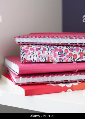 Stack of pink and floral pattern spiral notebooks, ring binders and notebooks on white shelf in home office, close-up Stock Photo