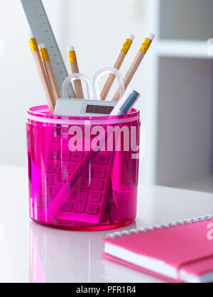 Pencils, ruler, pen, scissors and calculator in transparent pink plastic container on white desk next to pink spiral notebook in home office, close-up Stock Photo