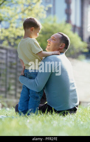 Man sitting down on grass, hugging boy around waist, looking at each other Stock Photo
