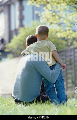 Man sitting on grass side by side with standing boy, hugging each other, view from behind Stock Photo