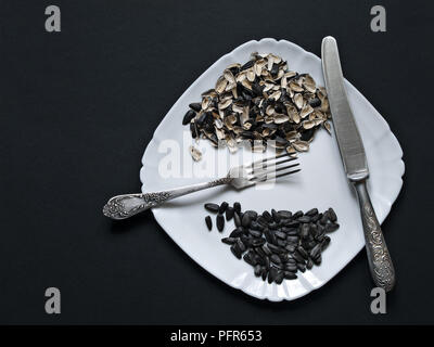 On a dark background there is a white plate with unrefined sunflower seeds, the husk of seeds, a knife, a fork shot from above with a close-up Stock Photo