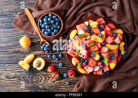 delicious colorful fresh fruit salad with watermelon, blueberries, peach slices, strawberry and lime on a clay plate on an old rustic table with ingre Stock Photo