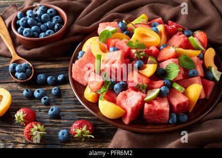 delicious colorful fresh fruit salad with watermelon, blueberries, peach slices, strawberry and lime on a clay plate on an old rustic table with ingre Stock Photo