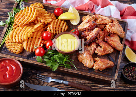 fried chicken wings, Crispy Potato Criss Cross Fries on a clay plates on a wooden table with mustard and tomato sauce dipping, view from above, close- Stock Photo