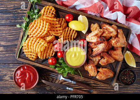 fried chicken wings, Crispy Potato Criss Cross Fries on a clay plates on a wooden table with mustard and tomato sauce dipping, view from above, close- Stock Photo