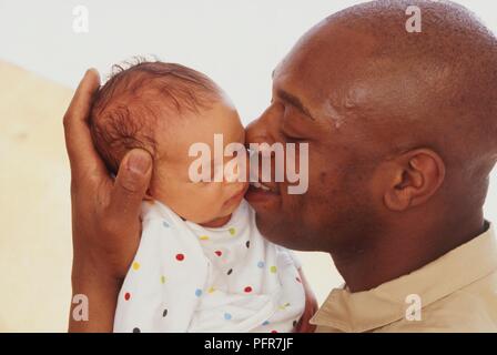 Man kissing young baby on the cheek, while supporting his head with his hand, side view Stock Photo