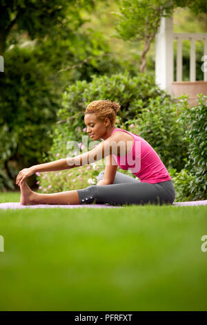 Young woman in leggings and pink top sitting in garden performing stretching exercise Stock Photo