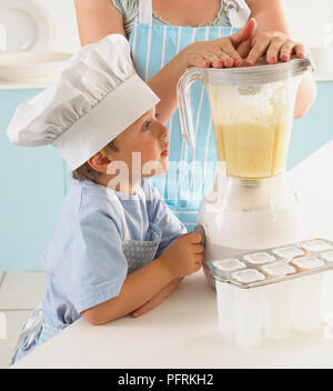 Boy in chef's hat turning on blender containing mixture of pineapple, sugar, coconut milk and lime, woman's hands holding down lid, mould for ice lollies on kitchen worktop Stock Photo