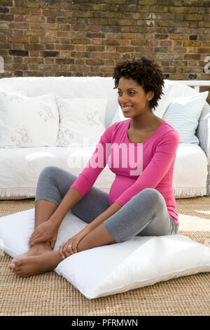 Young pregnant woman sitting on cushion pressing soles of feet together Stock Photo