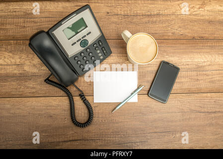 Top view of desk phone, blank note paper, pen, a cup of coffee and a smart phone, on a wooden table with copyspace. Stock Photo