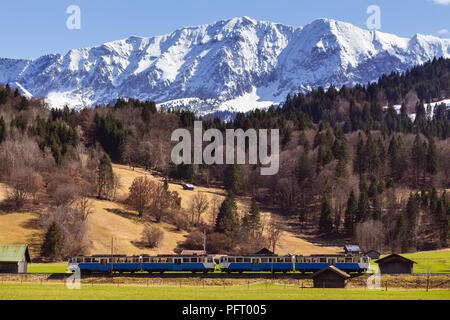 A train passes farm fields and barns near town of Garmisch Partenkirchen in Bavaria, Germany; forest and snow covered Alpes in background Stock Photo