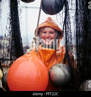 Rain in the 1960s. A young smiling woman is dressed in rain clothes and poses on a the deck of a fishing vessel with fishing nets. Sweden 1969 Stock Photo