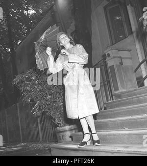 1940s woman in raincoat. A young woman dressed in a raincoat and holding an umbrella is looking up in the sky to see if the rain will keep pouring. Sweden 1946 ref V112-5 Stock Photo