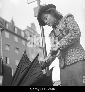 1940s woman in raincoat. Actress Viveca Lindfors, 1920-1995, is waiting at a bus stop and folds her umbrella together when it approaches. She is dressed in 1940s short chequered jacket and skirt, matching hat, handbag and gloves. August 1940. Photo Kristoffersson 159-20 Stock Photo