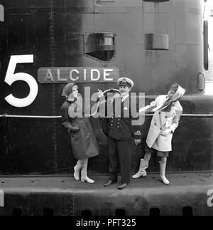 1960s fashion. Two fashion models and the captain of the British submarine Alcide. The submarine is anchored in the port of Stockholm. The models are wearing the autumns fashion of raincoats and head scarfs. Sweden May 1962 Stock Photo