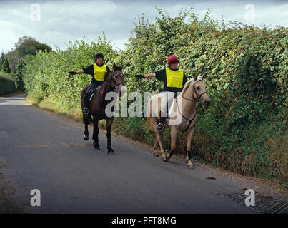 Two young women wearing fluorescent tops riding ponies on country lane using arms to signal right turn Stock Photo