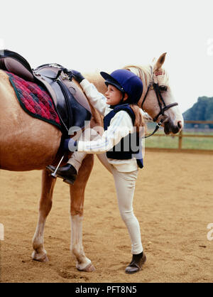 Young girl wearing riding habit, standing beside palomino pony, holding stirrup and saddle before mounting Stock Photo