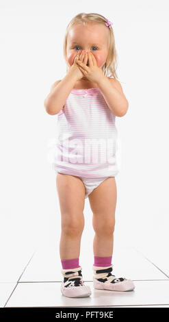 Girl wearing vest, pants and slippers, covering her face with her hands, 20 months Stock Photo