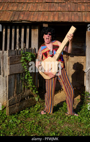 spoof portrait of older male wearing wig sunglasses bright clothing and medallion while playing a fake guitar mid life crisis concept hungary Stock Photo