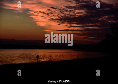 Boy fishing from the lakeshore in Villa Carlos Paz, Cordoba, Argentina, during a colorful cloudy sunset Stock Photo