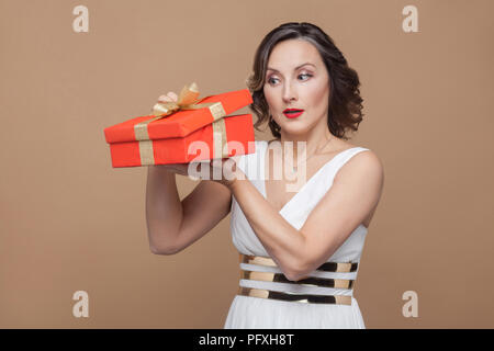 Cunning middle aged elegant brunette woman in white dress holding and looking inside the red gift box. Emotion and feeling concept. Studio shot, indoo Stock Photo