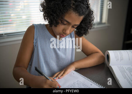 Young school girl studying at home at a table using a textbook and writing in a notebook Stock Photo