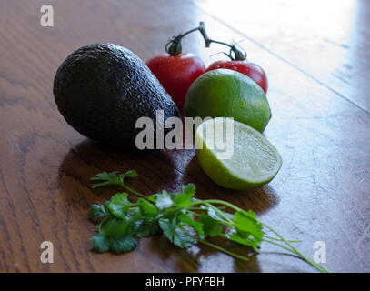 Avocado, lime cut in half, tomatoes on the vine and cilantro/koriander placed in a group on a wooden table with opposite light Stock Photo
