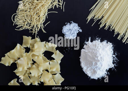Three different types of pasta, pile of flour, salt and pepper are laid out on black background Stock Photo