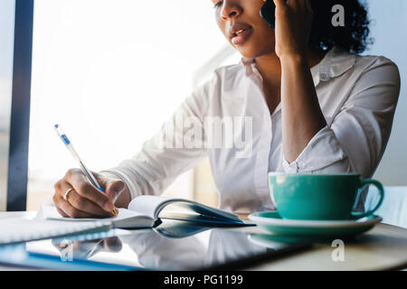 Close up of young entrepreneur woman taking notes while talking on mobile phone in a cafe Stock Photo