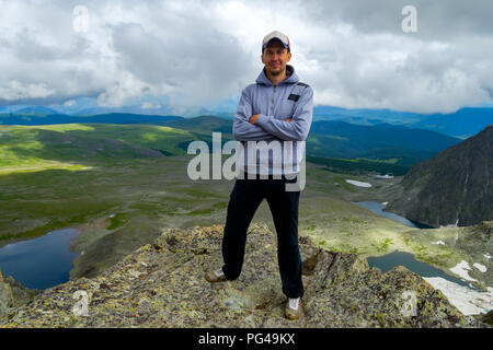 Man in a cap stands on the mountain with his hands clasped against the backdrop of a colorful valley landscape flooded with sunlight, hills, blue lake Stock Photo