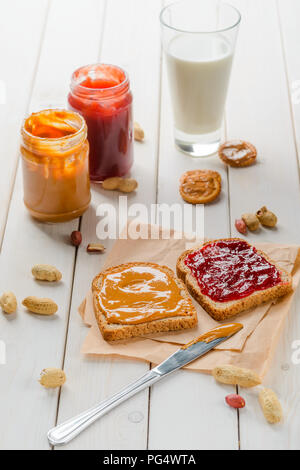 Yummy peanut butter and jam sandwich on baking paper. Jars, glass of milk, crackers. wooden background. Stock Photo