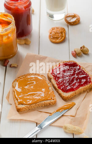 Traditional sandwich on baking paper, close-up. Peanut butter and jam jars, crackers and glass pf milk on the background. Stock Photo