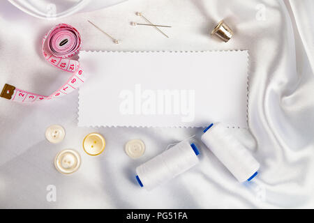 Sewing accessories and a note on white satin fabric. White satin fabric, thread, thimble, pins and measuring tape.