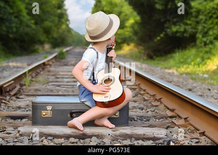 Boy playing guitar on tracks while sitting on a suitcase and waiting for a train in the sun during the summer. Stock Photo